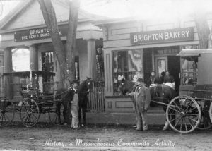 Photograph of the storefront Brighton Bakery and the Brighton Five Cents Saving Bank, with two horses and wagons, with their minders, in front of them.